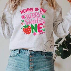 May include: A light gray crewneck sweatshirt featuring a graphic print with the text "MOMMY OF THE BERRY SWEET ONE" in pink and green. The design includes a red strawberry, small pink flowers, green leaves, and tiny red hearts.