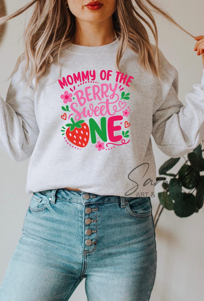 May include: A light gray crewneck sweatshirt featuring a graphic print with the text "MOMMY OF THE BERRY SWEET ONE" in pink and green. The design includes a red strawberry, small pink flowers, green leaves, and tiny red hearts.