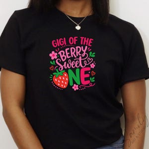 May include: A black short-sleeved t-shirt featuring a graphic print with the text GIGI OF THE BERRY SWEET ONE in pink and white lettering. The design includes a red strawberry, small pink flowers, green leaves, and tiny red hearts.