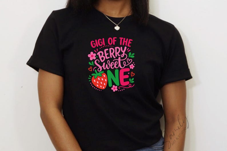 May include: A black short-sleeved t-shirt featuring a graphic print with the text GIGI OF THE BERRY SWEET ONE in pink and white lettering. The design includes a red strawberry, small pink flowers, green leaves, and tiny red hearts.