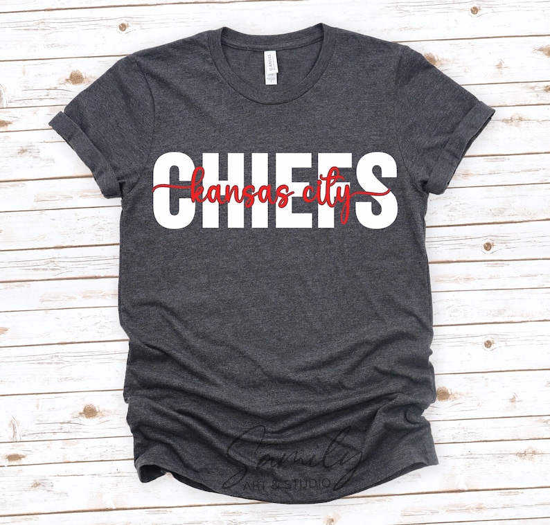 May include: Dark gray t-shirt with the word "CHIEFS" in large white block letters and "Kansas City" in red script. The shirt has rolled-up sleeves and a crew neck. The shirt is made of a soft material.