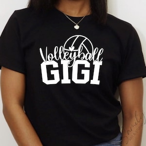May include: Black t-shirt featuring the text "Volleyball GIGI" in white, with a volleyball graphic. The shirt has short sleeves and is likely made of cotton. The text is in a mix of script and block letters.