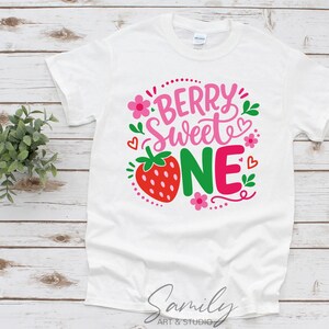 May include: A white short-sleeve t-shirt featuring a graphic print with the text "BERRY SWEET ONE" in pink and green. The design includes a red strawberry, small pink flowers, green leaves, and red heart outlines arranged around the lettering.