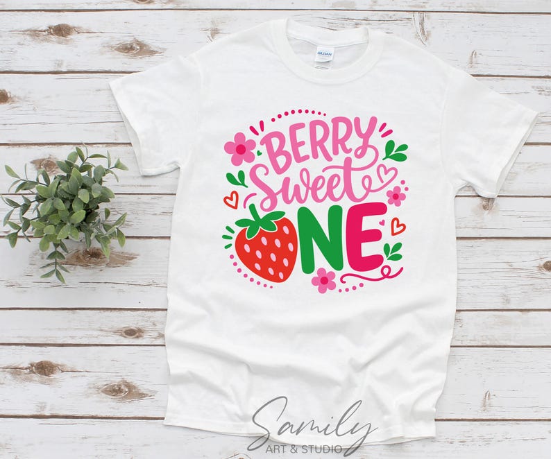 May include: A white short-sleeve t-shirt featuring a graphic print with the text "BERRY SWEET ONE" in pink and green. The design includes a red strawberry, small pink flowers, green leaves, and red heart outlines arranged around the lettering.