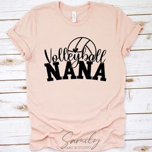May include: A pale pink t-shirt featuring the text "Volleyball Nana" in black, with a volleyball graphic. The shirt is presented on a light wood-effect surface.