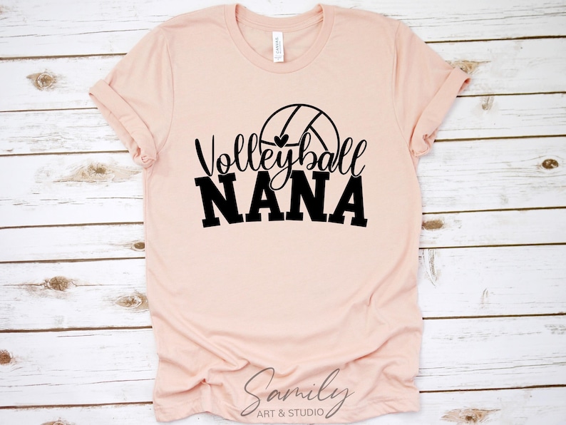 May include: A pale pink t-shirt featuring the text "Volleyball Nana" in black, with a volleyball graphic. The shirt is presented on a light wood-effect surface.