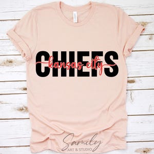 May include: A light pink t-shirt with the word "CHIEFS" in large black letters, with "Kansas City" in a red script font underneath. The shirt is made of soft material and has rolled-up sleeves.