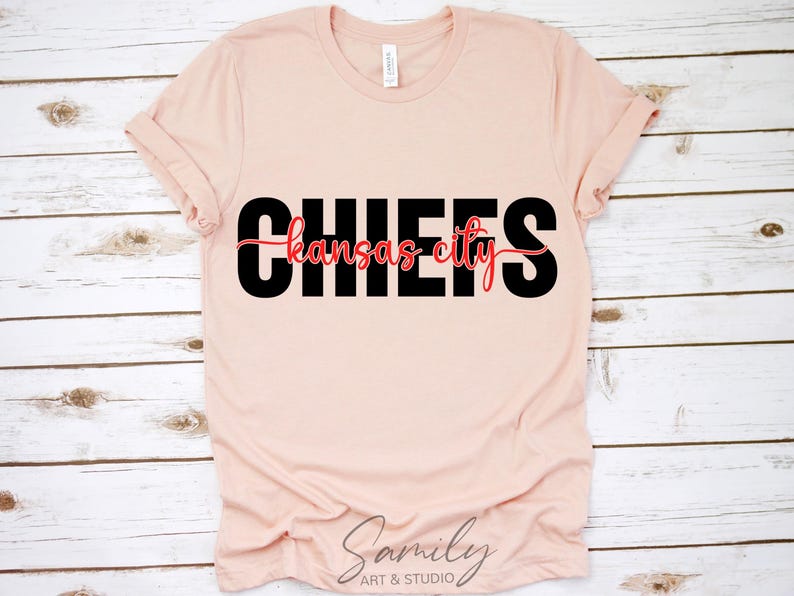 May include: A light pink t-shirt with the word "CHIEFS" in large black letters, with "Kansas City" in a red script font underneath. The shirt is made of soft material and has rolled-up sleeves.