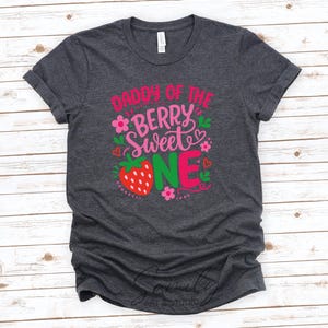 May include: A dark heather gray short-sleeve t-shirt featuring the text DADDY OF THE BERRY SWEET ONE in pink, red, and green. The design includes a red strawberry graphic, small pink flowers, and tiny red hearts printed on the front center.