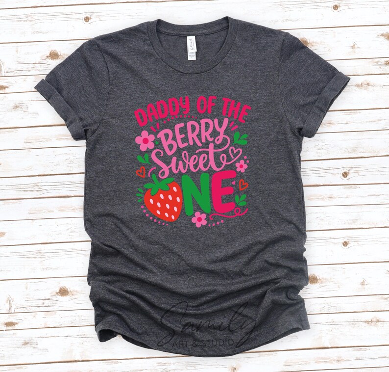 May include: A dark heather gray short-sleeve t-shirt featuring the text DADDY OF THE BERRY SWEET ONE in pink, red, and green. The design includes a red strawberry graphic, small pink flowers, and tiny red hearts printed on the front center.