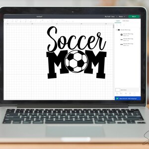May include: A laptop displays a black and white graphic that reads "Soccer Mom" with a soccer ball in the center. The laptop is open on a wooden surface, with the keyboard visible. The screen shows a design program interface.