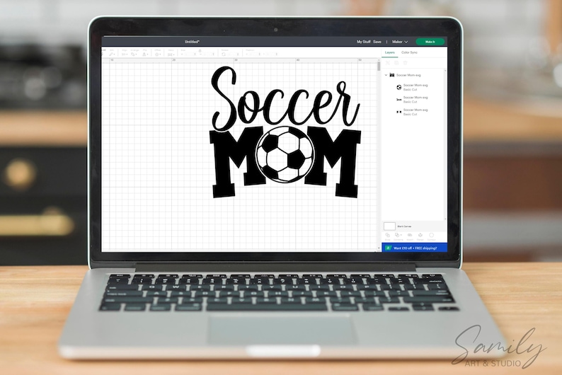 May include: A laptop displays a black and white graphic that reads "Soccer Mom" with a soccer ball in the center. The laptop is open on a wooden surface, with the keyboard visible. The screen shows a design program interface.