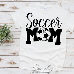 May include: White t-shirt with the words "Soccer MOM" in black script and block letters. A soccer ball graphic with the number "20" is in the center. The shirt is on a light wood surface.