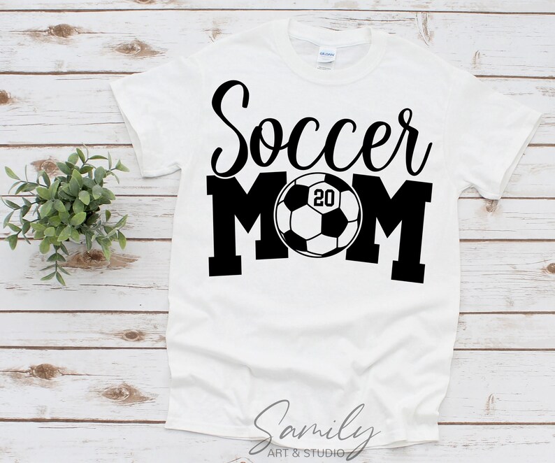 May include: White t-shirt with the words "Soccer MOM" in black script and block letters. A soccer ball graphic with the number "20" is in the center. The shirt is on a light wood surface.