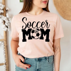 May include: A light pink t-shirt with the words "Soccer Mom" in black script and block letters. A soccer ball graphic with the number "20" is in the center. The shirt is short-sleeved and made of a soft material.