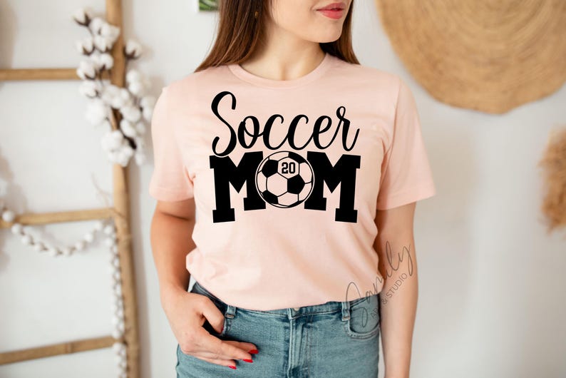 May include: A light pink t-shirt with the words "Soccer Mom" in black script and block letters. A soccer ball graphic with the number "20" is in the center. The shirt is short-sleeved and made of a soft material.