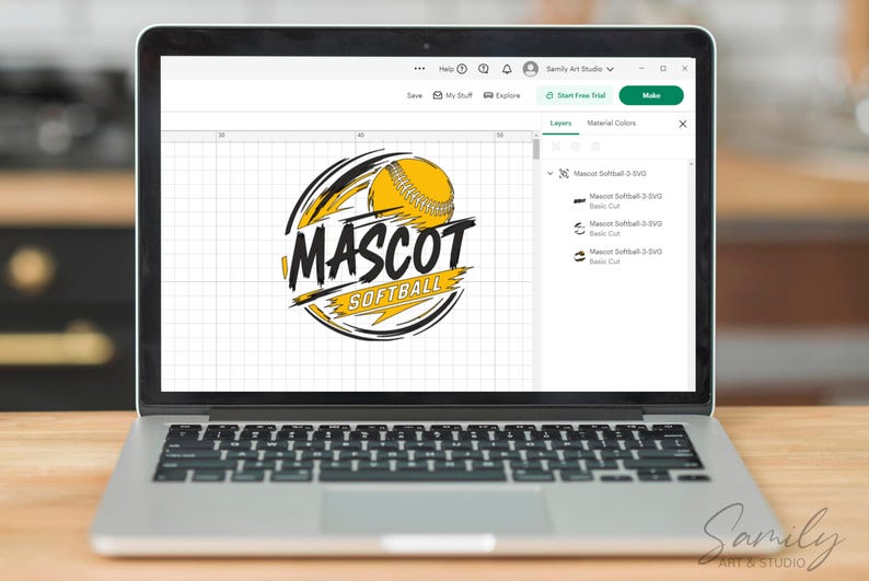 May include: Laptop displaying a softball logo design. The logo features a yellow softball with black and yellow accents, and the words "MASCOT SOFTBALL" in bold, stylized text. The laptop is on a wooden surface.