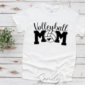 May include: White t-shirt with the words "Volleyball Mom" in black script and block letters. The "O" in "Mom" is a volleyball with a heart. The shirt is on a light wood surface.