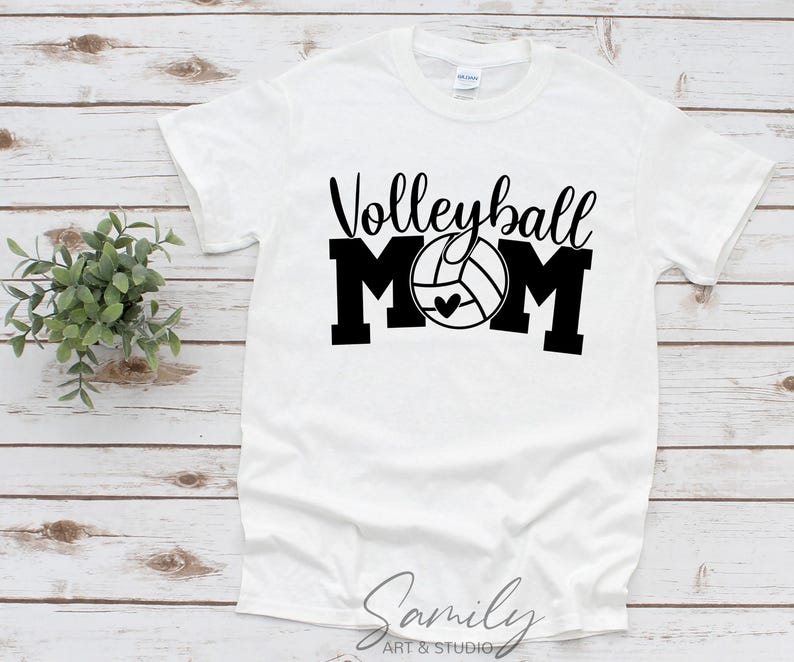 May include: White t-shirt with the words "Volleyball Mom" in black script and block letters. The "O" in "Mom" is a volleyball with a heart. The shirt is on a light wood surface.