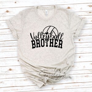 May include: A light beige t-shirt featuring the text "Volleyball Brother" in black script and block letters, with a volleyball graphic. The shirt is made of a soft material and has rolled-up sleeves.