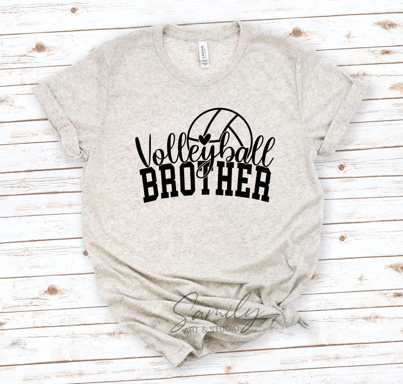 May include: A light beige t-shirt featuring the text "Volleyball Brother" in black script and block letters, with a volleyball graphic. The shirt is made of a soft material and has rolled-up sleeves.