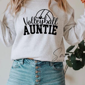 May include: A light grey sweatshirt featuring the text "Volleyball Auntie" in black, with a volleyball graphic. The sweatshirt is worn with light blue jeans. The image is a lifestyle shot, likely for apparel.