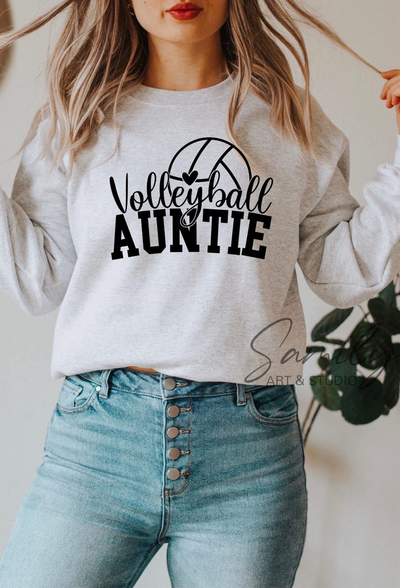 May include: A light grey sweatshirt featuring the text "Volleyball Auntie" in black, with a volleyball graphic. The sweatshirt is worn with light blue jeans. The image is a lifestyle shot, likely for apparel.