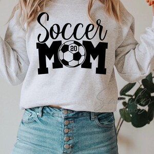 May include: A light gray sweatshirt with the words "Soccer MOM" in black script and block letters, with a soccer ball graphic in the center. The soccer ball has the number "20" inside. The person is wearing blue jeans.
