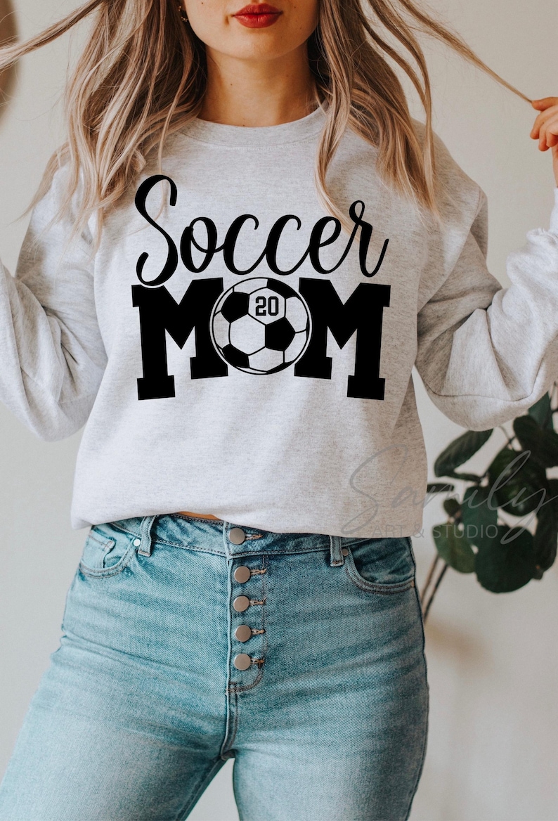 May include: A light gray sweatshirt with the words "Soccer MOM" in black script and block letters, with a soccer ball graphic in the center. The soccer ball has the number "20" inside. The person is wearing blue jeans.