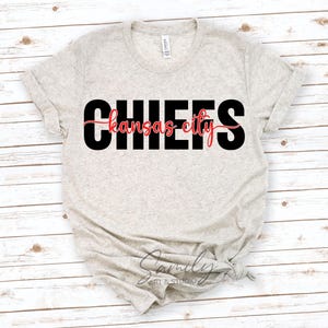 May include: A light beige t-shirt with the word "CHIEFS" in large black block letters. Below, in red script, is the text "kansas city". The shirt is knotted at the bottom and has a tag that reads "Canvas".