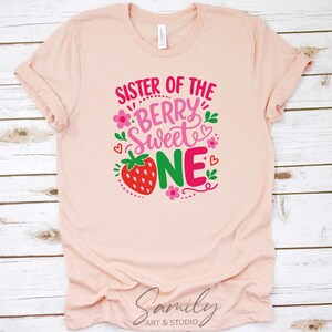 May include: A light peach-colored short-sleeve t-shirt featuring a graphic print with the text "SISTER OF THE BERRY SWEET ONE" in pink and red. The design includes a red strawberry, small pink flowers, green leaves, and red hearts.