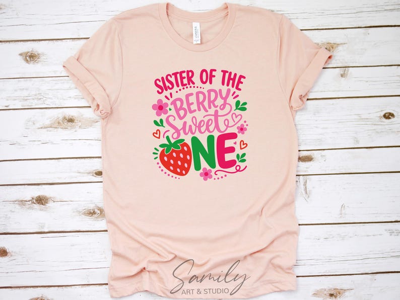 May include: A light peach-colored short-sleeve t-shirt featuring a graphic print with the text "SISTER OF THE BERRY SWEET ONE" in pink and red. The design includes a red strawberry, small pink flowers, green leaves, and red hearts.