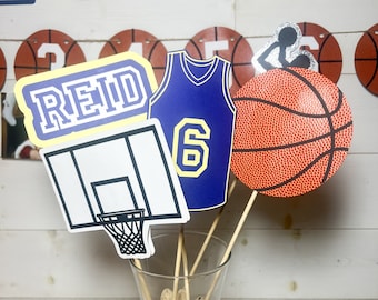 May include: A basketball themed centerpiece with a basketball, a basketball jersey with the number 6, a basketball hoop, and a sign that says "Reid". The items are placed in a clear glass vase filled with peanuts.