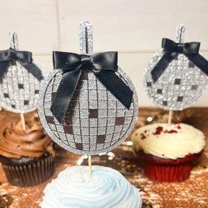 Puede incluir: Tres cupcakes con adornos de bola de discoteca plateada con purpurina y lazos negros. Los cupcakes están cubiertos con glaseado blanco, marrón y rojo. Los cupcakes están sobre una superficie de lentejuelas doradas.