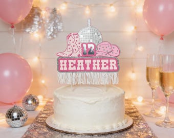 May include: A white frosted cake with a decorative topper featuring a disco ball, cowboy boots, and a hat. The topper has the name "HEATHER" and the number "12" in pink and silver glitter. Pink balloons and disco balls are in the background.