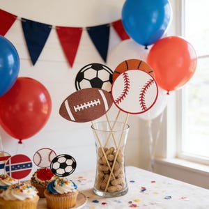 Può includere: Un'esposizione festosa a tema sportivo. Un vaso di vetro trasparente contiene arachidi, con ritagli di football americano, baseball e calcio su bastoncini. Palloncini rossi, bianchi e blu e uno striscione decorano lo sfondo. Cupcake con decorazioni sportive completano la scena.