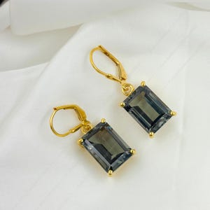 May include: A pair of gold-colored leverback earrings. Each earring features a rectangular, smoky gray gemstone. The gemstones are set in gold-colored prongs at each corner. The earrings are displayed on a white, textured surface.
