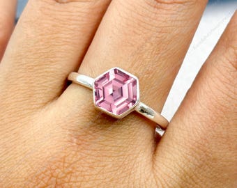May include: A silver ring featuring a hexagonal, light pink gemstone. The gemstone is set in a bezel setting and has a faceted design. The ring is shown on a person's finger.