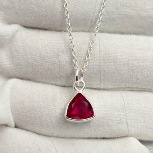 Puede incluir: Un collar de plata con un colgante triangular de piedra preciosa rojo intenso. El colgante está engastado en un bisel de plata y cuelga de una delicada cadena de plata. La piedra preciosa tiene una superficie facetada.
