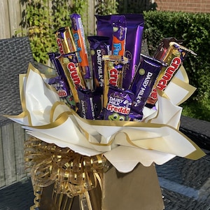 May include: A gift arrangement featuring various Cadbury chocolate bars including Dairy Milk, Crunchie, and Freddo, displayed in a gold-coloured box. The chocolates are wrapped in white and gold paper, accented with a large gold ribbon and curly gold ribbons.