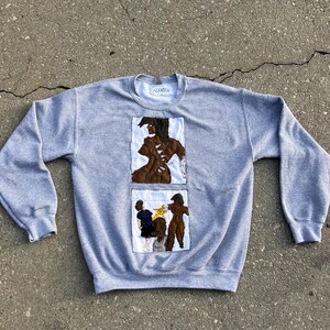 May include: A gray sweatshirt with two embroidered patches. The top patch features a brown figure on a white background. The bottom patch shows two figures and a yellow object. The sweatshirt has a crew neck.