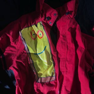 Peut inclure: Veste rouge à capuche avec un motif rectangulaire jaune sur le devant. Le rectangle jaune représente un visage de dessin animé avec des cercles rouges pour les yeux et un sourire bleu. La veste a une fermeture éclair et des détails noirs.