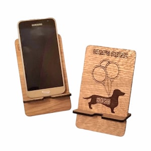 May include: Two wooden phone stands featuring a dachshund design. The stand on the right has the text "SAM'S PHONE" and "DASH" on it. The stand on the left has a black smartphone in it.