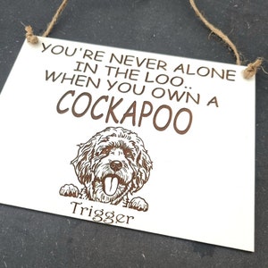 May include: White wooden sign with brown text that reads "You're never alone in the loo when you own a Cockapoo" with a cartoon image of a Cockapoo dog peeking over the top of the sign. The dog's name "Trigger" is written below the image.