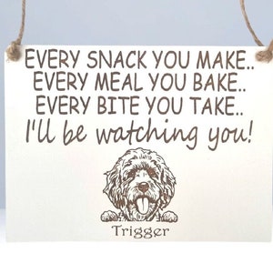 May include: A white wooden sign with brown text that reads "EVERY SNACK YOU MAKE.. EVERY MEAL YOU BAKE.. EVERY BITE YOU TAKE.. I'll be watching you!"  A cartoon image of a dog with the name "Trigger" is below the text.