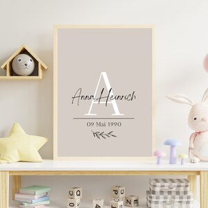 Könnte beinhalten: Gerahmter Druck mit dem Namen "Anna Heinrich" und dem Datum "09 Mai 1990" in einer stilvollen Schriftart. Der Druck zeigt einen großen Buchstaben "A" und einen Zweig mit Blättern. Der Druck wird auf einem weißen Regal mit Spielzeug ausgestellt.