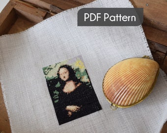 May include: A white Aida cloth with a cross-stitch pattern of the Mona Lisa painting. The cloth is in a wooden box with a seashell on top of the cloth. The text "PDF Pattern" is on a black rectangle.