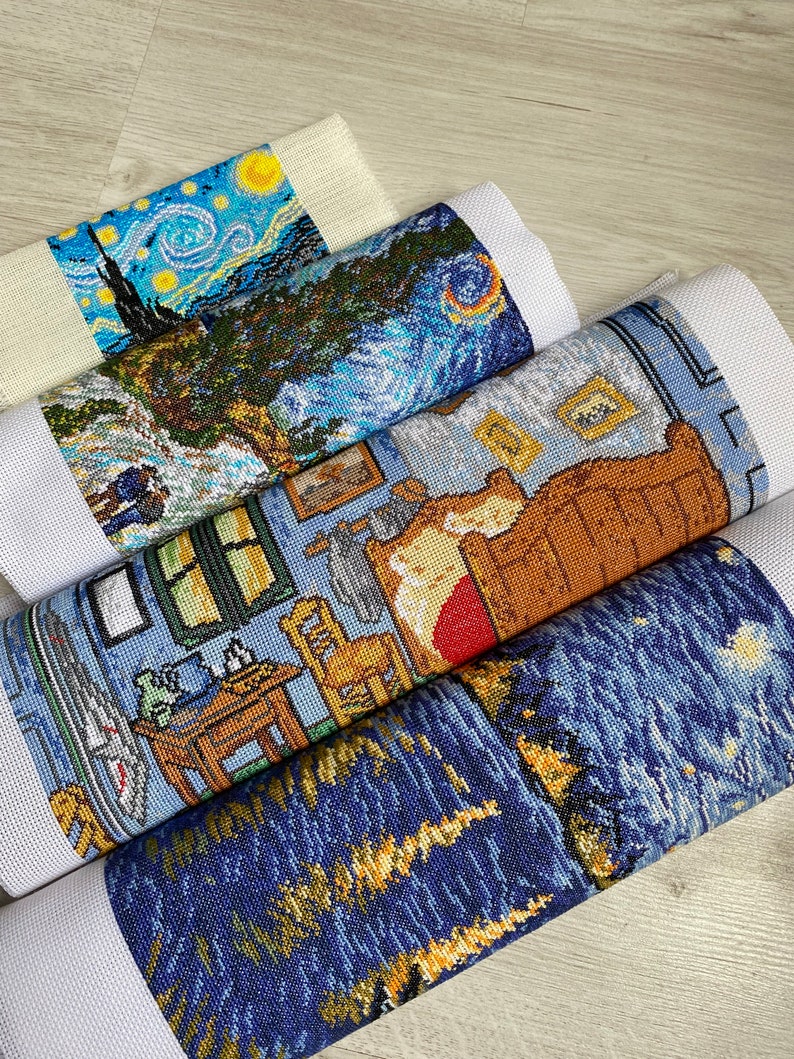 Op de afbeelding: Vier opgerolde stukken Aida-doek met kruissteekborduursels van beroemde schilderijen. Het bovenste stuk is een blauwe en gele sterrenhemel-sc&egrave;ne, het tweede is een blauw en groen landschap, het derde is een bruine en blauwe slaapkamer-sc&egrave;ne en het onderste is een andere blauwe en gele sterrenhemel-sc&egrave;ne.