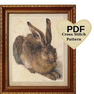 May include: A framed cross-stitch pattern of a brown hare with long ears, set against a cream-coloured background. The artwork is in a decorative, ornate, dark brown wooden frame. A heart-shaped tag with the text "PDF Cross Stitch Pattern" is on the right.