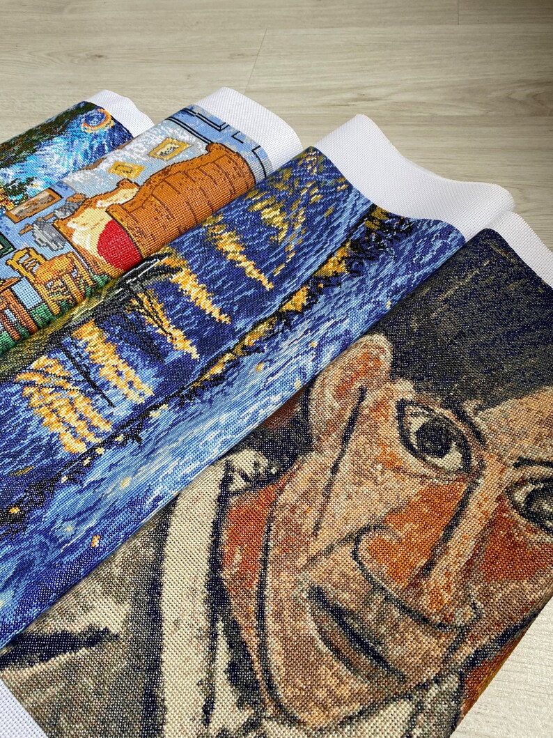 Op de afbeelding: Een stapel van vier onafgewerkte kruissteek-borduurprojecten. Het bovenste project is een blauwe en gele sterrenhemel-sc&egrave;ne. Het tweede project is een blauwe en gele sc&egrave;ne met een gebouw en een boom. Het derde project is een bruine en gele sc&egrave;ne met een vat. Het onderste project is een close-up van een gezicht met bruine huid en donker haar.